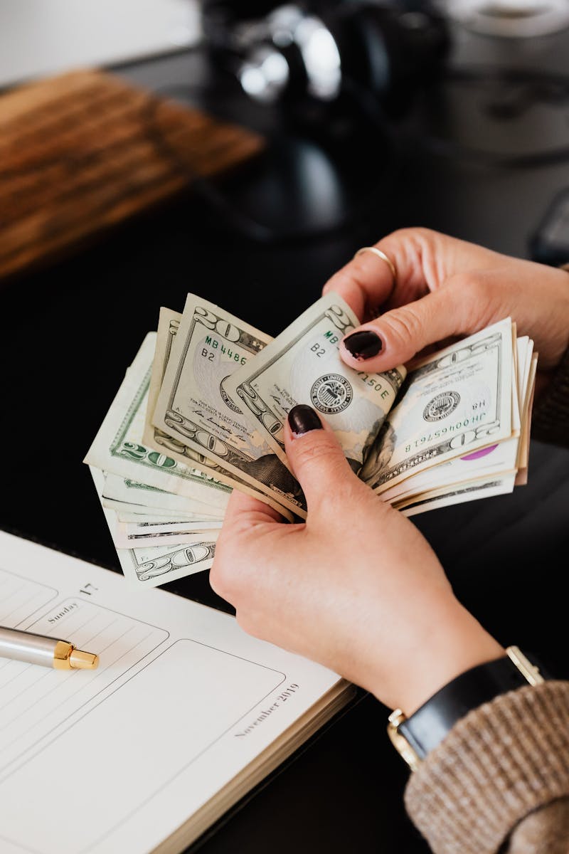 Unrecognizable elegant female in sweater counting dollar bills while sitting at wooden table with planner and pen
